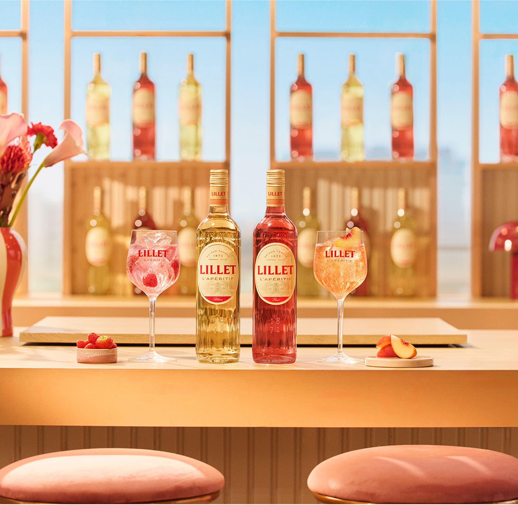 Zwei Flaschen Lillet Blanc und Lillet Rosé mit sommerlichen Aperitif-Drinks auf einer Bar vor dekorativem Flaschenregal.
