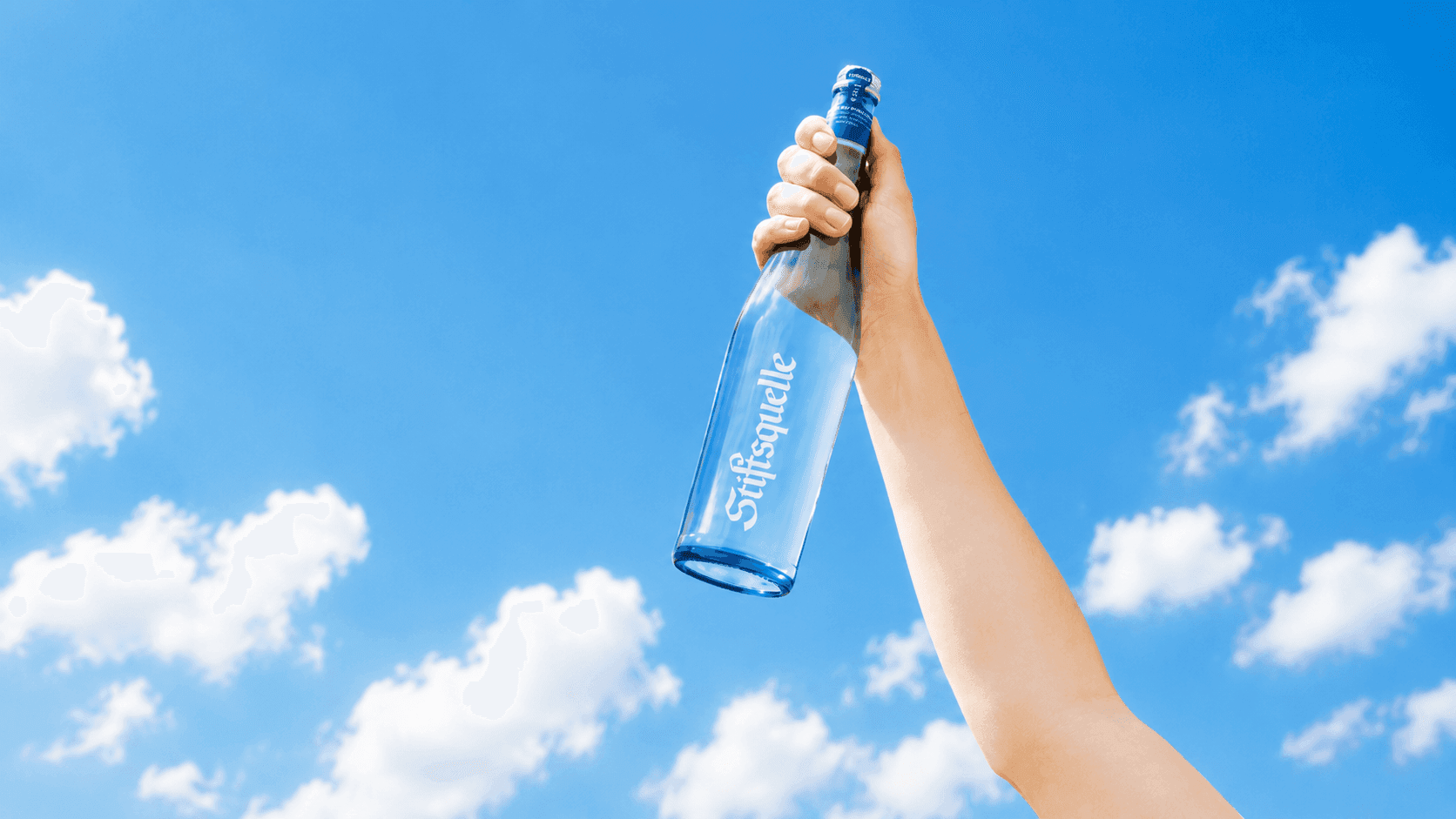 Hand hält eine Stiftsquelle-Glasflasche vor blauem Himmel mit weißen Wolken.